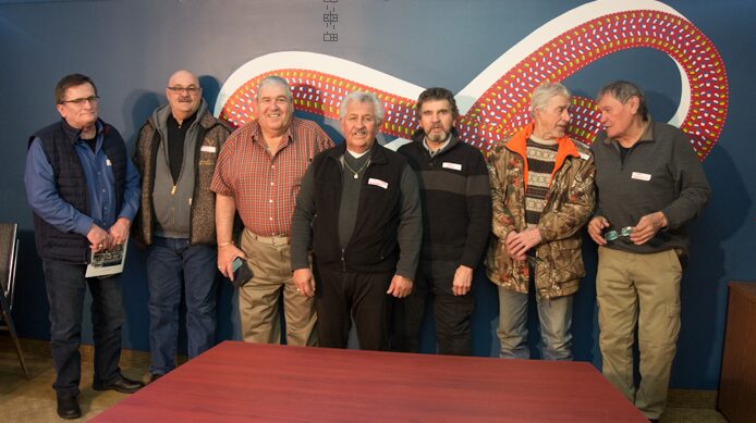 Seven older men standing in an office in front of a wall with a red Metis symbol.