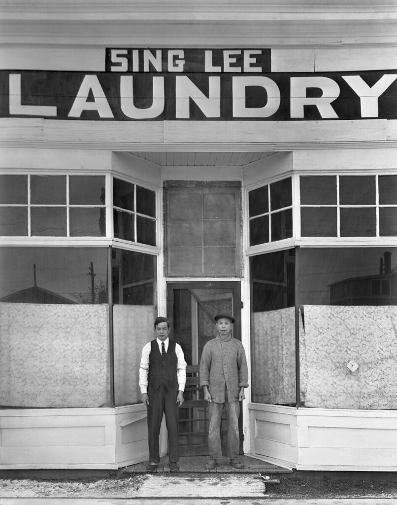 Two men in front of a storefront with the sign Sing Lee Laundry above them. They appear to be Chinese. The lower half of the store windows are papered over.