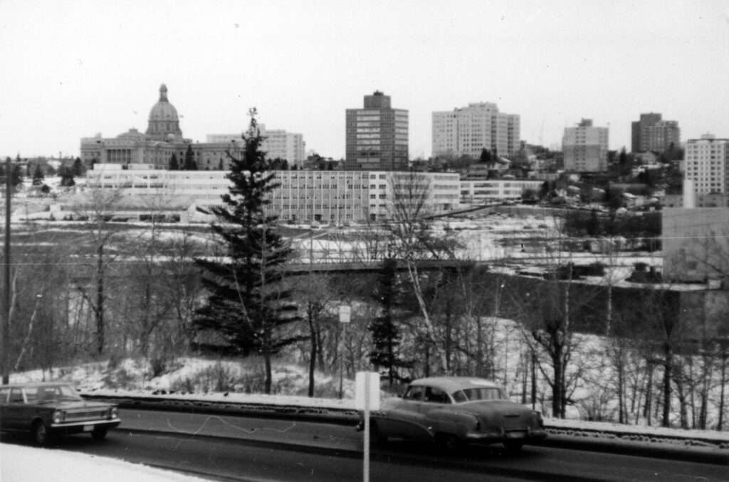 A long white rectangular building almost at river level, in front of the domed Alberta Legislature and several modest skyscrapers. In the middle of the scene is the Walterdale Bridge. In the foreground, cars drive down a road.