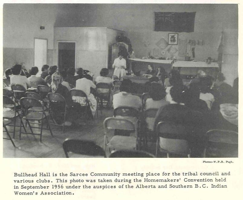 A classroom or hall with many metal folding chairs set out. A couple dozen women are seated watching a woman at the front of the room, who is reading something aloud. Behind her, we can see a variety of items, including a portrait of a man with a feathered hat, the flag of the dominion, and various textiles.