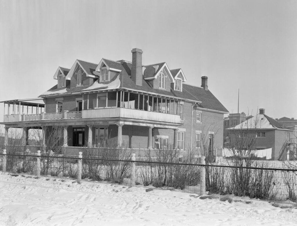 A large three-story brick house, pictured on a snowy day, with wraparound porches on the first and second floors. There is also a small brick stable house behind the main building.