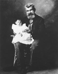 Photograph. A heavily bearded man in a black suit holds a toddler dressed in white on his knee.