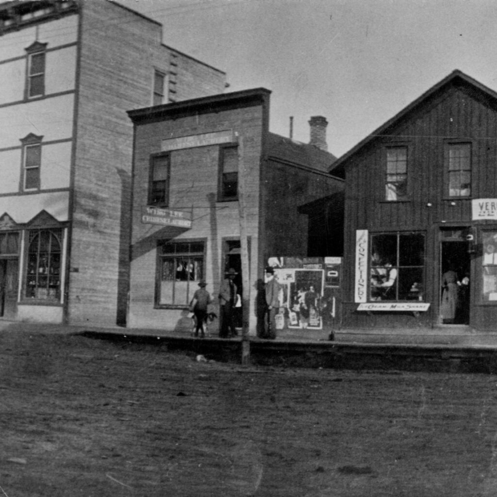 A black and white photograph of a streetscape on Jasper Avenue. There is a dirt road, and a few one and two story tall wooden buildings. The middle building has a sign that reads Wing Lee Chinese Laundry. Three people in suits and hats are standing near the door.