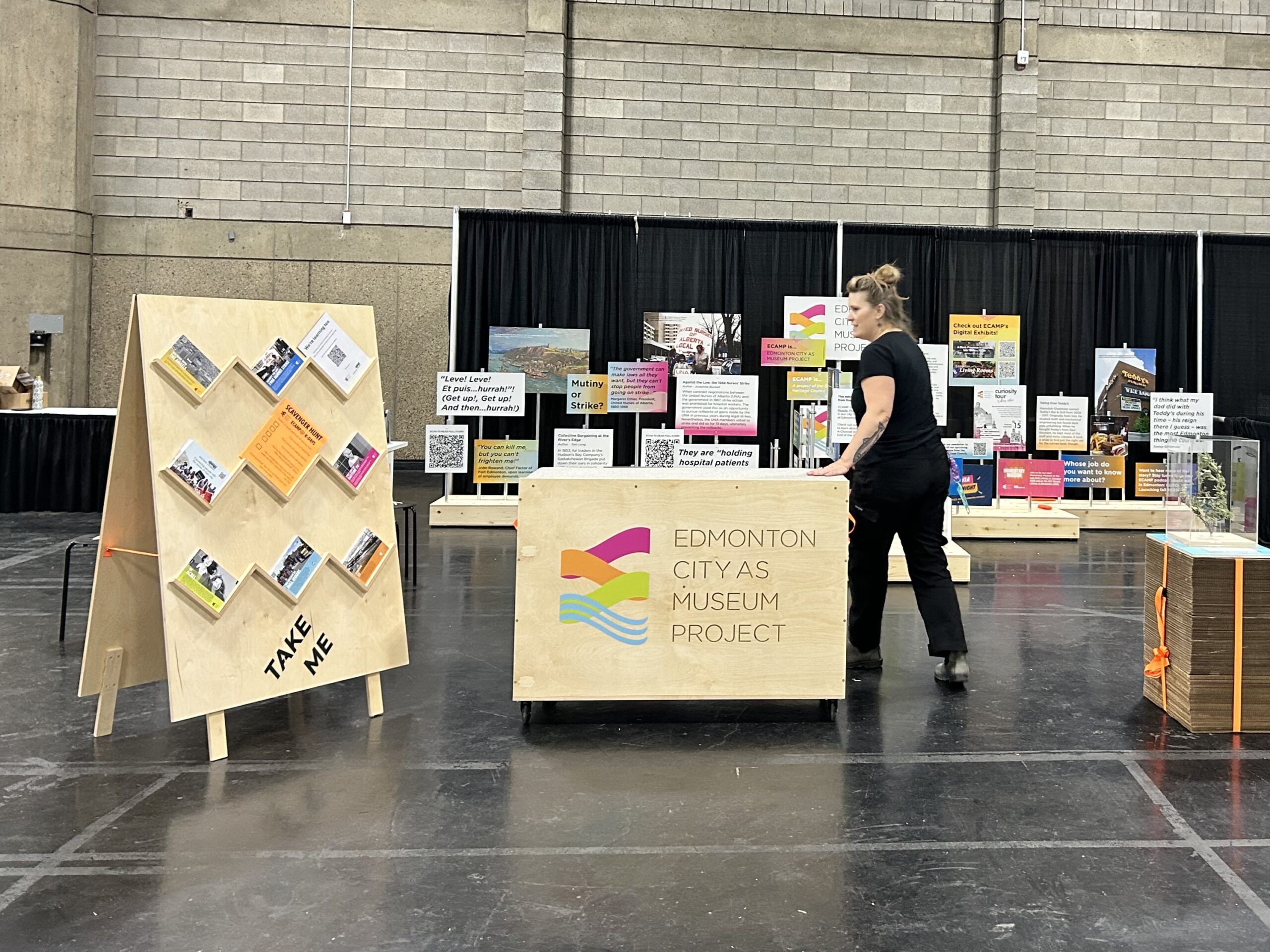 The ECAMP exhibit in Hall B. In the background, a couple dozen exhibit panels look like a rainbow of protest signs. In front, ECAMP Exhibit Coordinator Marina Hulzenga stands by a wooden cart with the ECAMP logo. There is a big sandwich board with many postcards, and it says TAKE ME.