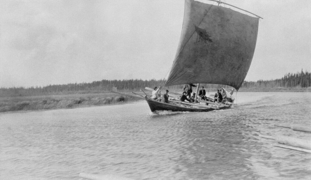 A wooden boat crewed by several men with oars leans to one side as a square sail full of wind propels it along a river.