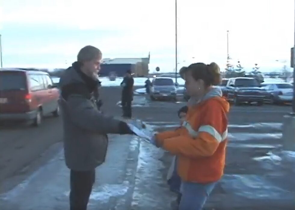 A man handing a pamphlet to a woman with a child in a snowy parking lot.