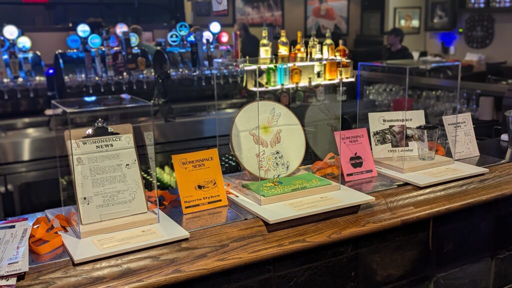 A series of clear display cases on a bar, including magazines, a t-shirt, and a calendar.