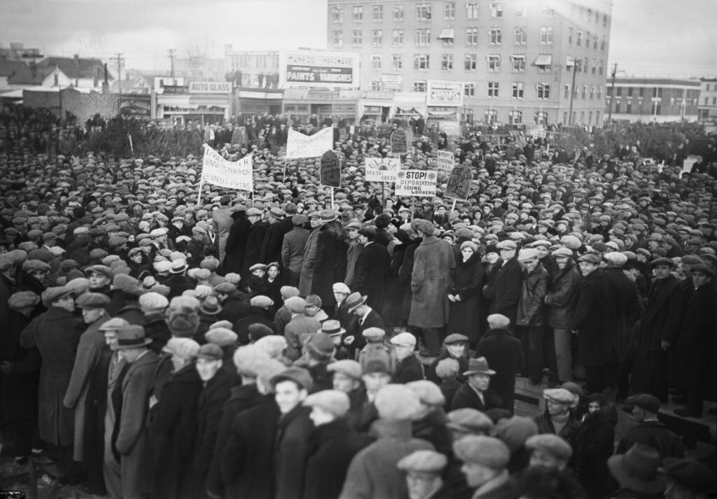 A black and white photograph of thousands of people downtown in an Edmonton square. Some hold protest signs with slogans like Stop Deportation of Young Workers and Our Living is First