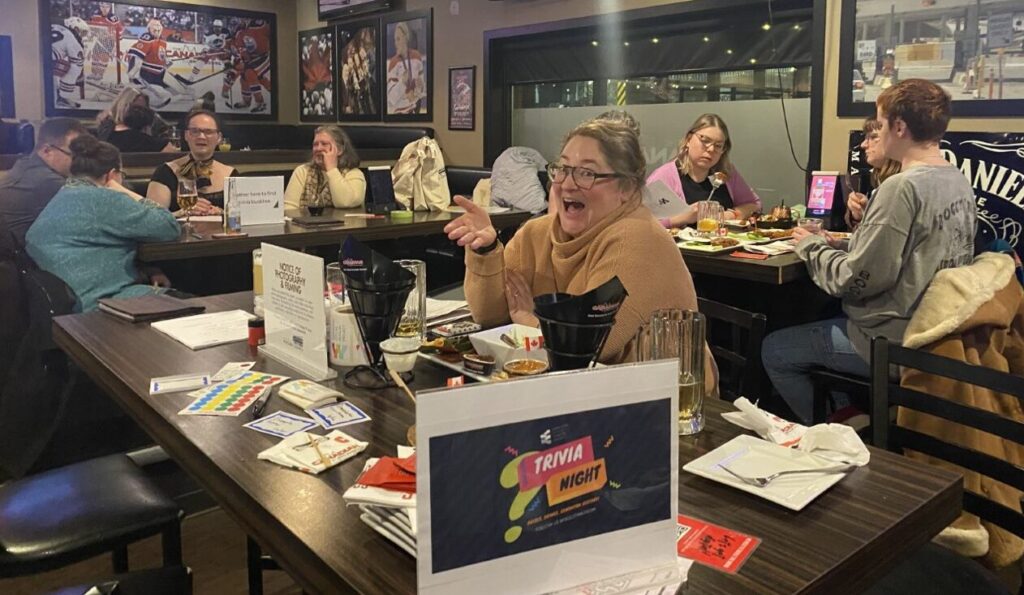 A smiling person at a table in a bar, with a crowd of patrons behind them.