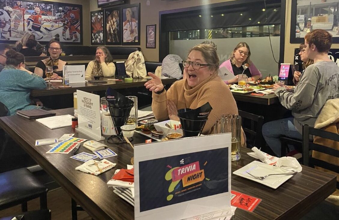 A smiling person at a table in a bar, with a crowd of patrons behind them.