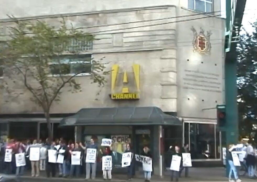 A photograph of workers with picket signs standing outside the Bay Building downtown, with the A-Channel "A" logo above them.